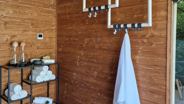 Un interior de madera con un perchero, repisas con toallas, una mesita con artículos de tocador y una planta en maceta.