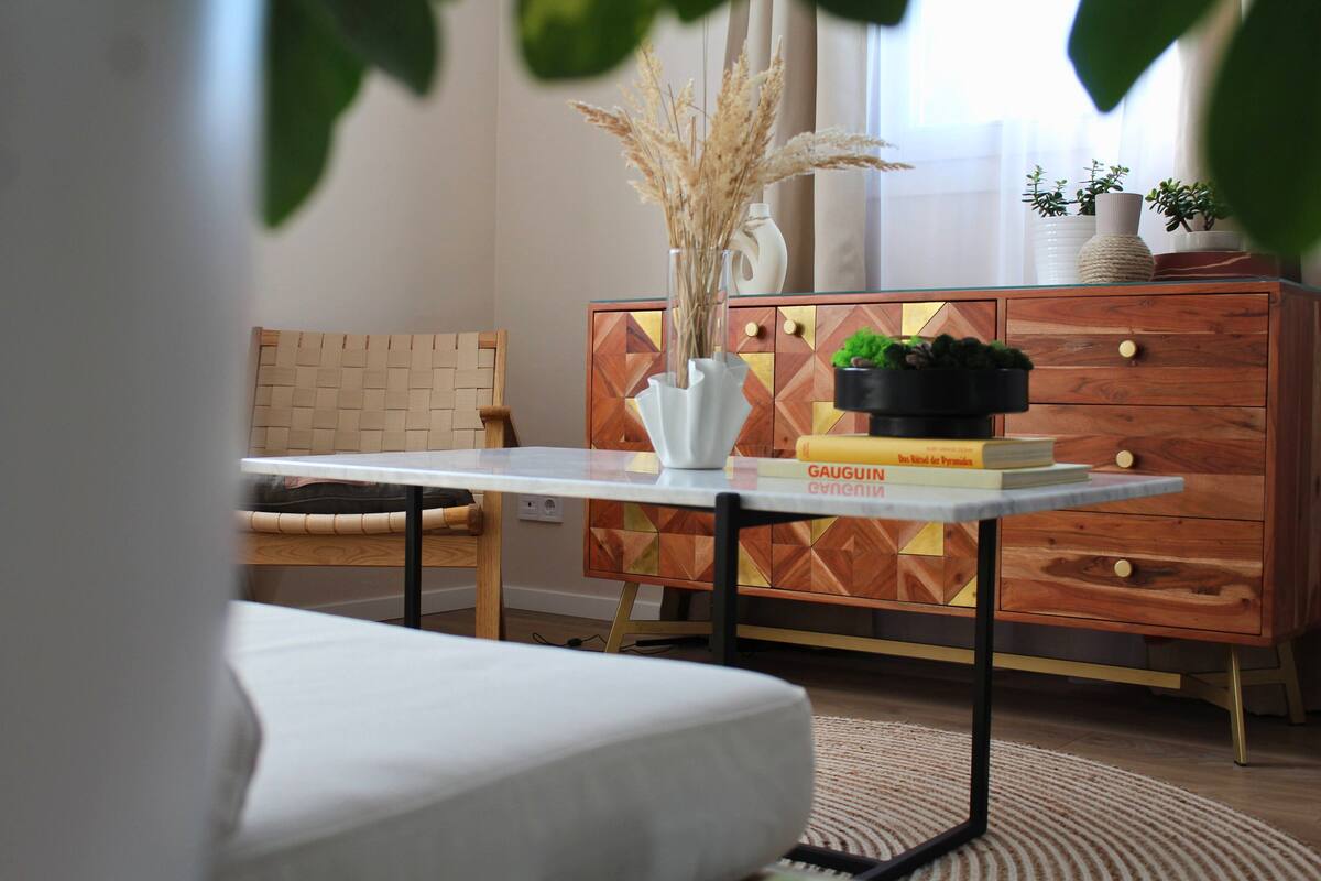 A modern living room with a white sofa, a wooden cabinet with geometric patterns, a small table with a vase of dried plants, and a potted plant on the cabinet.