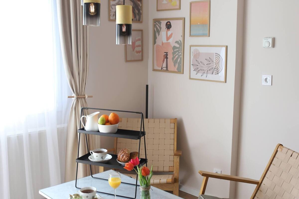 A modern living room with a round woven rug, a glass coffee table, a wicker chair, and a three-tiered side table with fruits and a teapot.