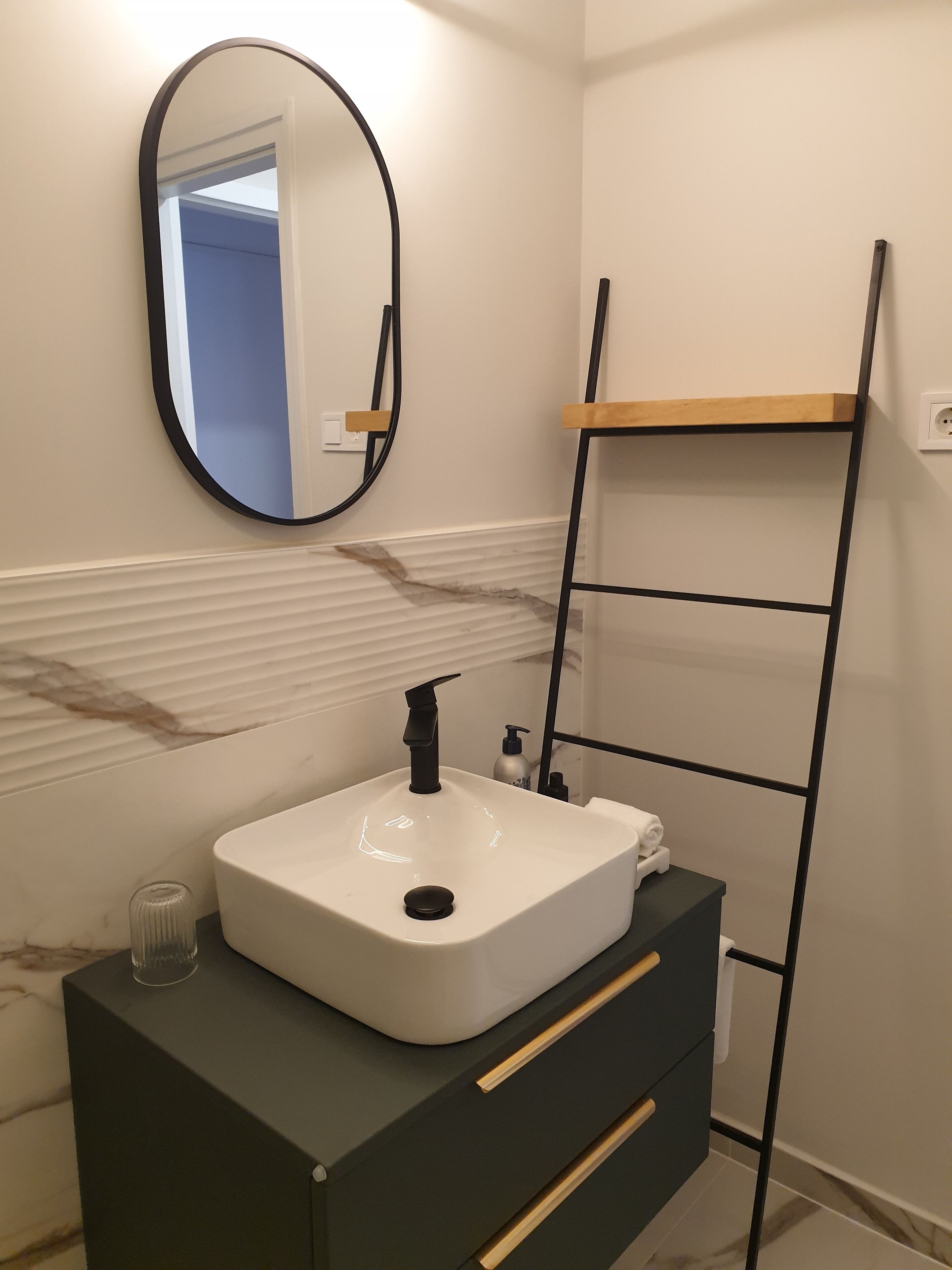 A bathroom with a white sink, black faucet, and a mirror. There is a black ladder leaning against the wall, and a wooden shelf above the sink.