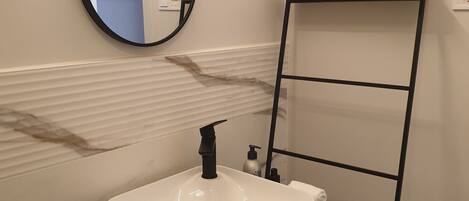 A bathroom with a white sink, black faucet, and a mirror. There is a black ladder leaning against the wall, and a wooden shelf above the sink.