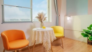 Une pièce avec une grande fenêtre, une petite table ronde recouverte d’une nappe en dentelle blanche, une chaise orange, une chaise jaune et une plante en pot.