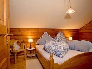 A cozy bedroom with a wooden headboard, a bed with floral bedding, two bedside lamps, and a small round table with a chair. - Mitterdorf Modern Retreat (Philippsreut)