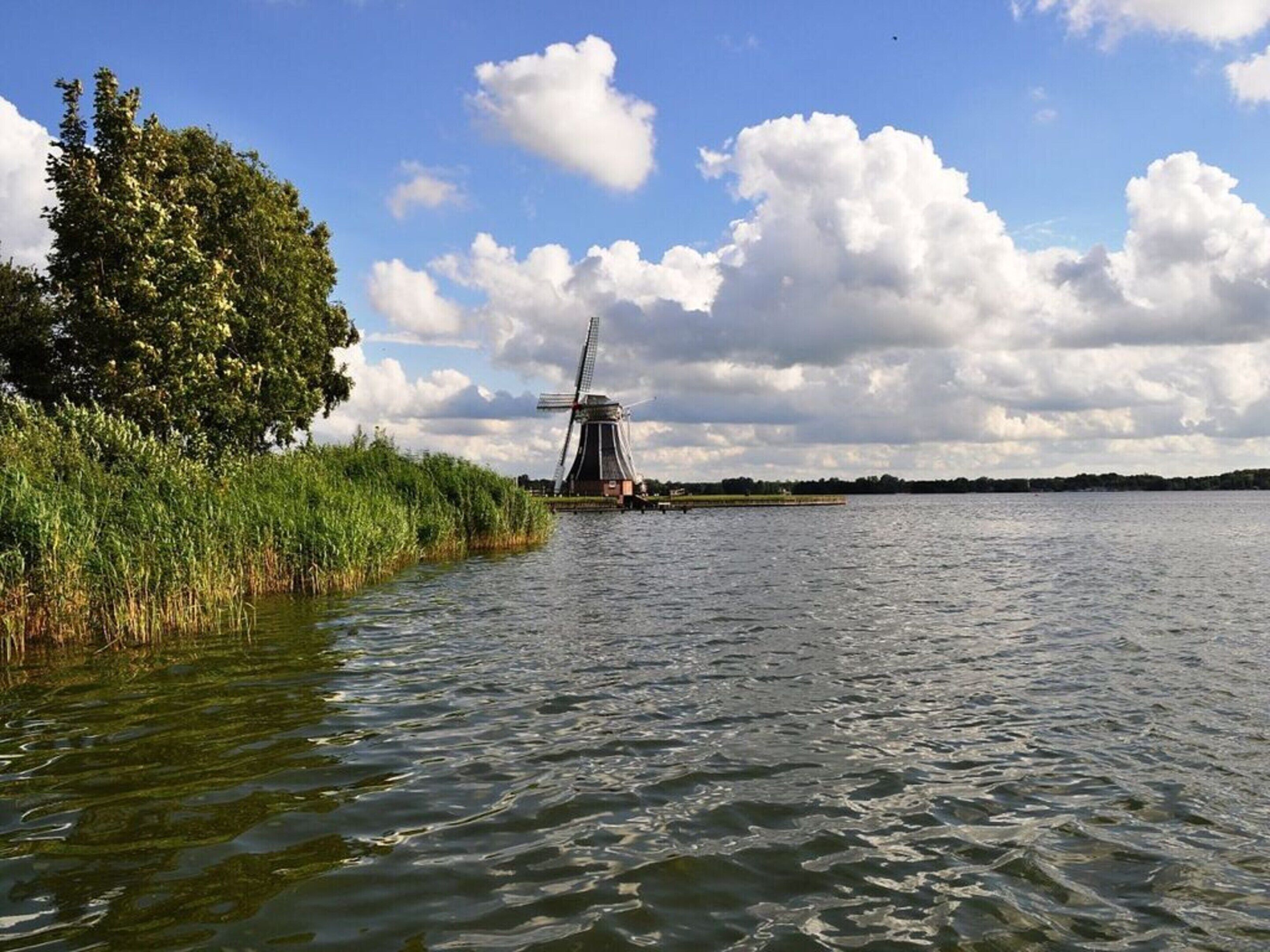 A windmill by a calm water body with a tree-lined shore.