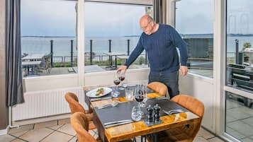 Et spiseområde med et bord dækket til to personer, store vinduer der giver udsigt over havet, og en mand, der hælder vin.