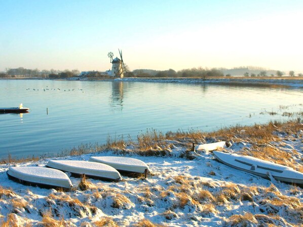 Un paysage paisible au bord d’un lac, avec un moulin à vent à l’horizon, un sol recouvert de neige et des bateaux sur la rive.
