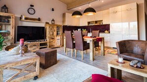 Un vaste espace de vie doté d’un plafond en bois, d’un téléviseur, d’une table à manger avec des chaises, et d’un coin cuisine avec des placards et des luminaires suspendus.