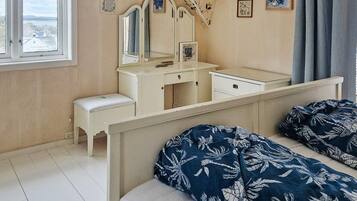 A bedroom with a white dresser, a vanity with mirrors, a bed with a floral bedspread, and a window with a view of the sea.