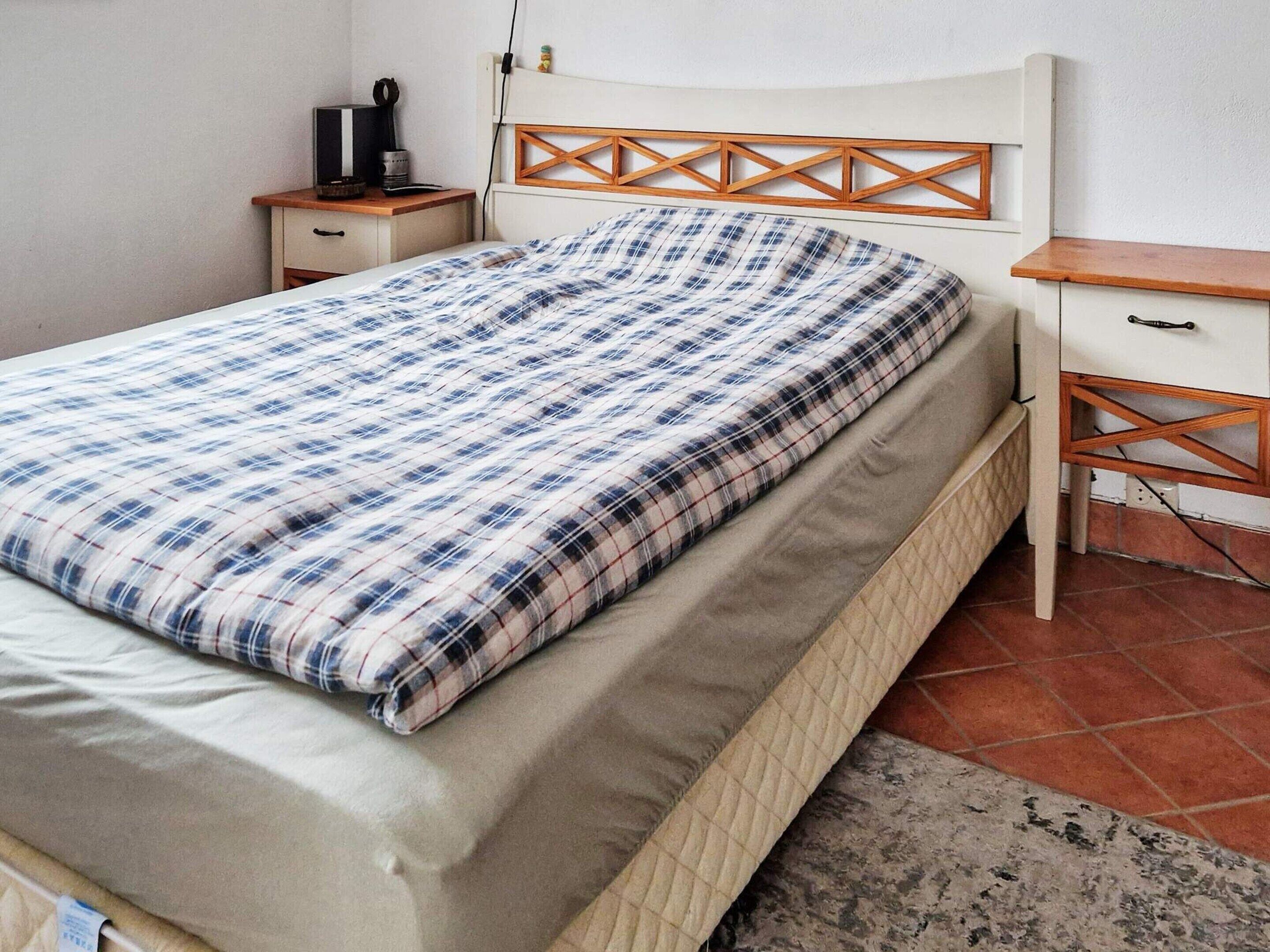 A neatly made bed with a plaid bedspread, a wooden headboard, a bedside table with a lamp, and a small drawer unit.