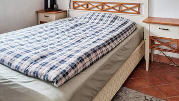 A neatly made bed with a plaid bedspread, a wooden headboard, a bedside table with a lamp, and a small drawer unit.