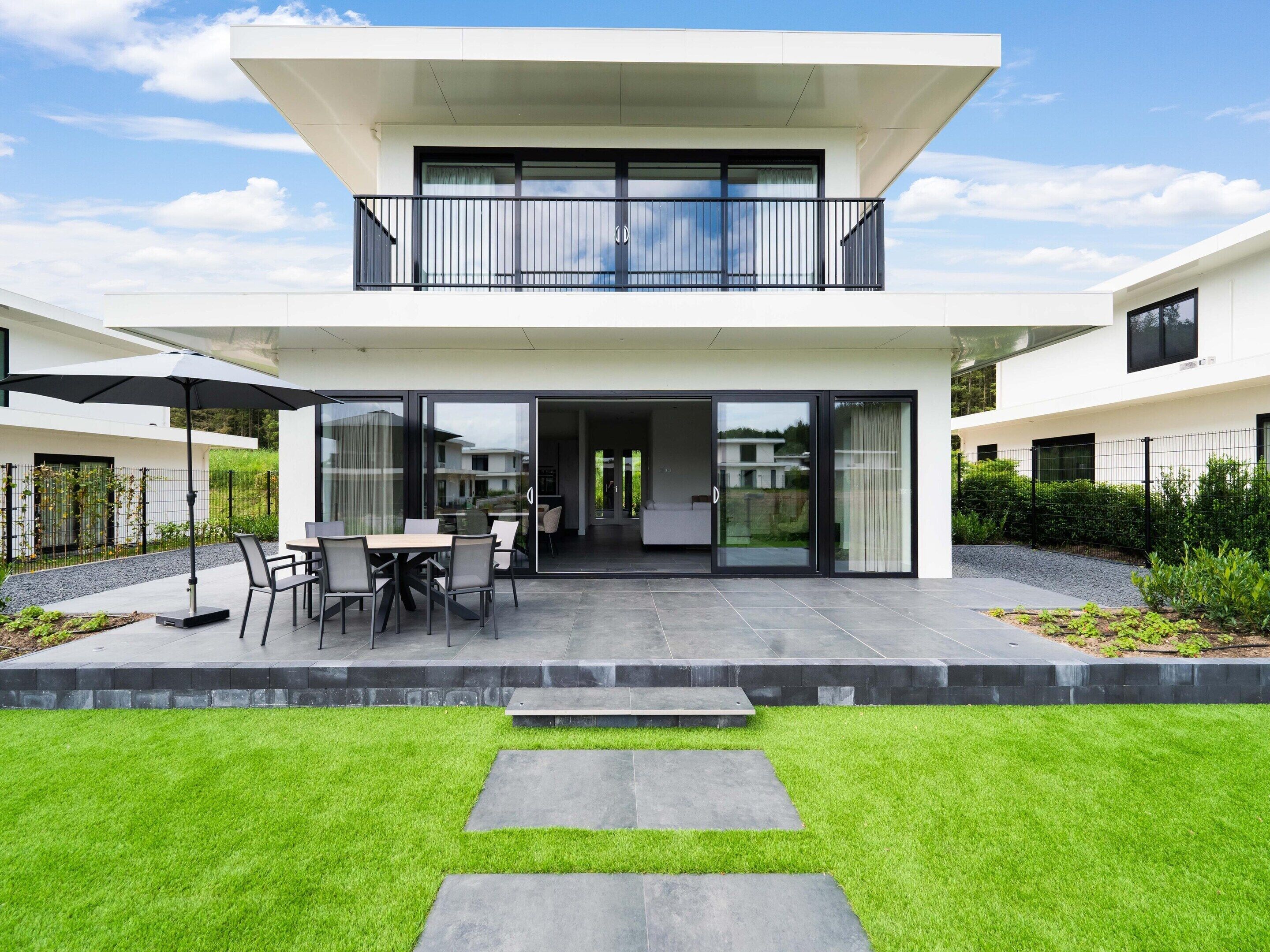 Ein modernes, zweistöckiges Haus mit Balkon, Terrasse und Gartenmöbeln sowie einem gepflegten Rasen.