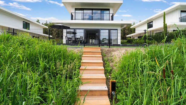 Een modern gebouw met twee verdiepingen, met een centrale ingang, omringd door hoog gras en een houten pad dat naar de ingang leidt.