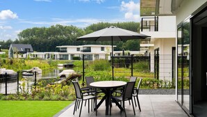 Een terras met een ronde tafel en stoelen, een zwarte parasol, en een uitzicht op een rivier en huizen op de achtergrond.
