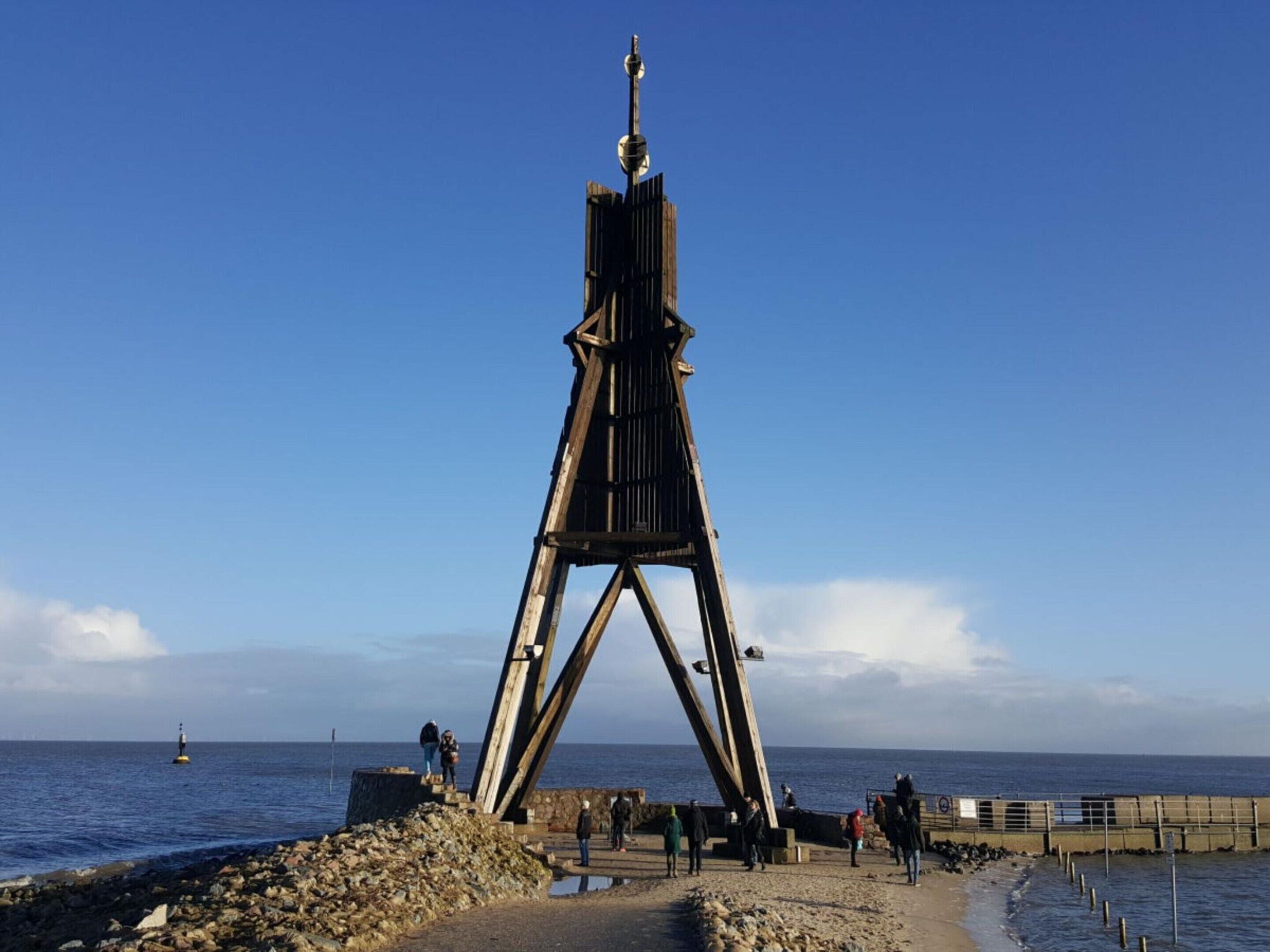 A lighthouse structure with a wooden base and a metal top, situated on a rocky shoreline with a clear view of the sea.