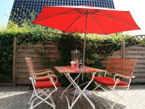 A patio with a wooden table and two chairs, an umbrella, and a brick wall with greenery. - Holiday Home Nige Og (Cuxhaven)