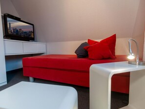 A small, modern room with a red sofa, a white desk, and a TV displaying a cityscape. - Holiday Home Nige Og (Cuxhaven)
