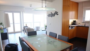 A modern kitchen with wooden cabinets, a dining area with a glass table, and a view through the glass door. - Apartment in Nieuwpoort With sea View (Nieuwpoort)