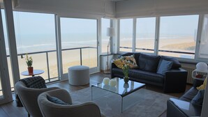 A spacious living room with a large glass table, comfortable seating, and a view of the beach. - Apartment in Nieuwpoort With sea View (Nieuwpoort)