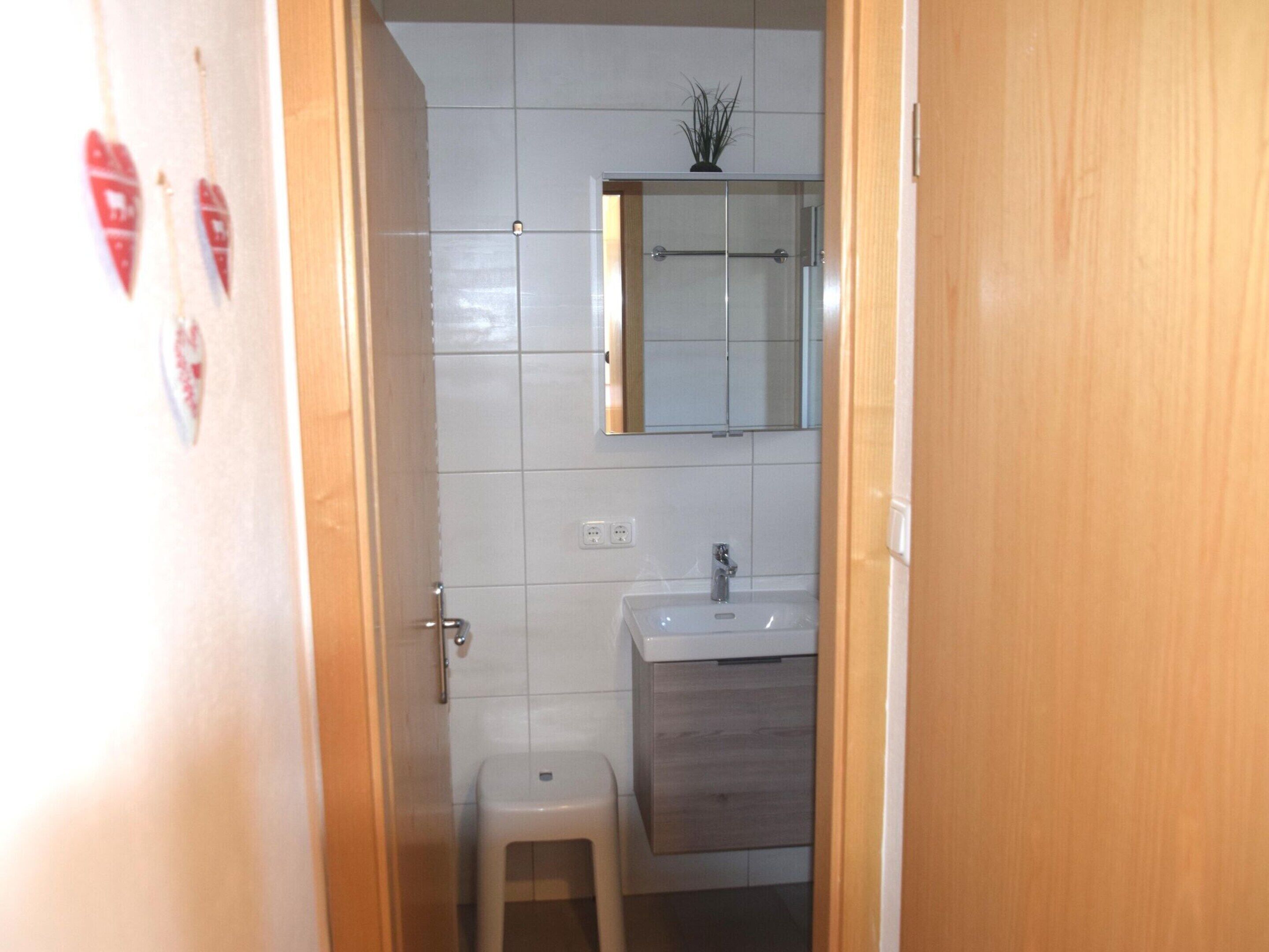A small bathroom with white tiles, a wooden door, a mirror, a sink, and a small stool.