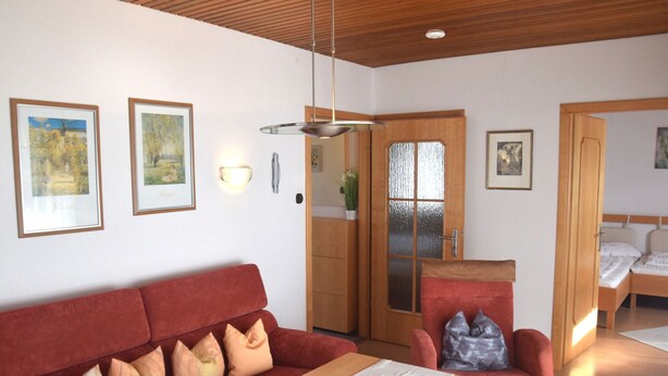 A modern room with a red sofa, a wooden table, and a bed with two pillows. There are framed pictures on the wall and a door with frosted glass. - Gemutlicher Fussener Ruckzugsort in der Nahe des Sees (Fuessen)