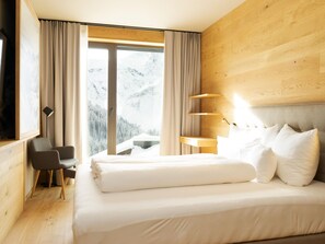 Hotelkamer met een bed, bureau, stoel en uitzicht op besneeuwde bergen door grote ramen.