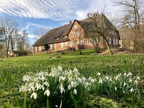 A large brick house with a thatched roof, surrounded by a grassy field and snowdrops. - Nordseelodge Comfortable Holiday Residence (Tetenbüll)