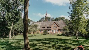 A traditional house with a thatched roof, surrounded by a lush green lawn and trees. - Nordseelodge Comfortable Holiday Residence (Tetenbüll)