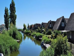 A row of traditional houses with thatched roofs, a calm water body in the foreground, and greenery surrounding the area. - Beautiful Villa With Winter Garden (Makkum)