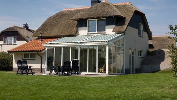 Une maison avec un toit de chaume, une véranda en verre et un jardin agrémenté d’un espace salon extérieur.