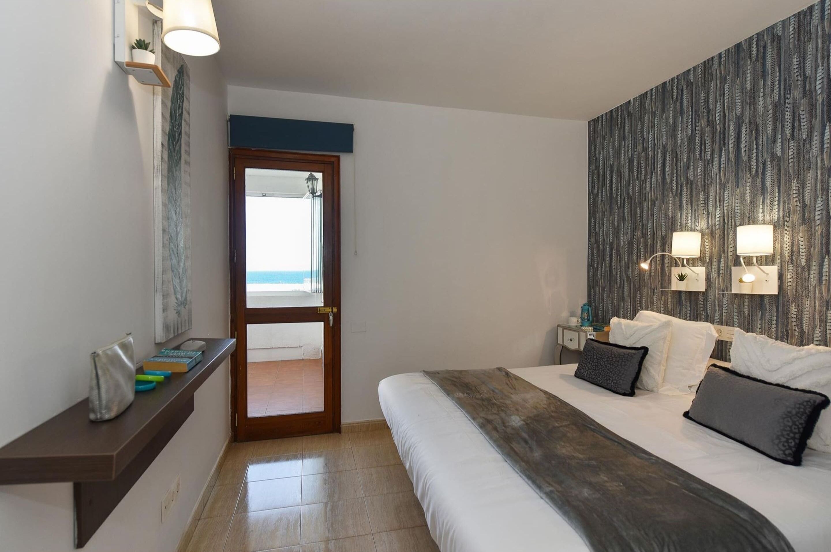A hotel room with a bed, bedside lamps, a nightstand, a shelf with books and a bag, and a view of the beach through a door.