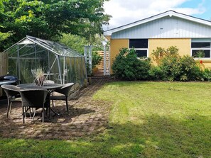 A greenhouse with a table and chairs, a building with a yellow exterior, and a grassy yard.