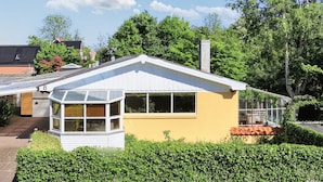 Een gelijkvloers huis met een tuinkamer, een zichtbare schoorsteen en een verzorgde haag ervoor.