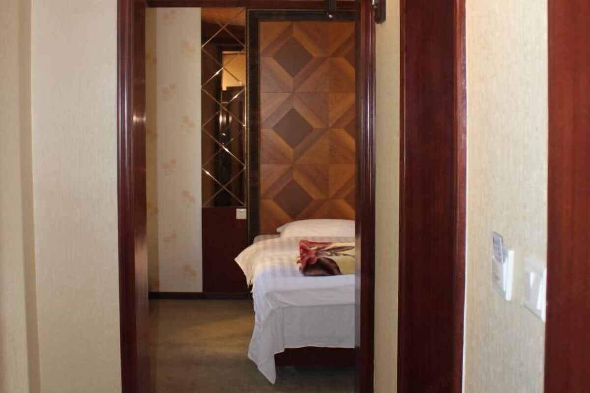 A narrow hallway leading to a room with a bed, a patterned wall, and a door with a wooden frame.