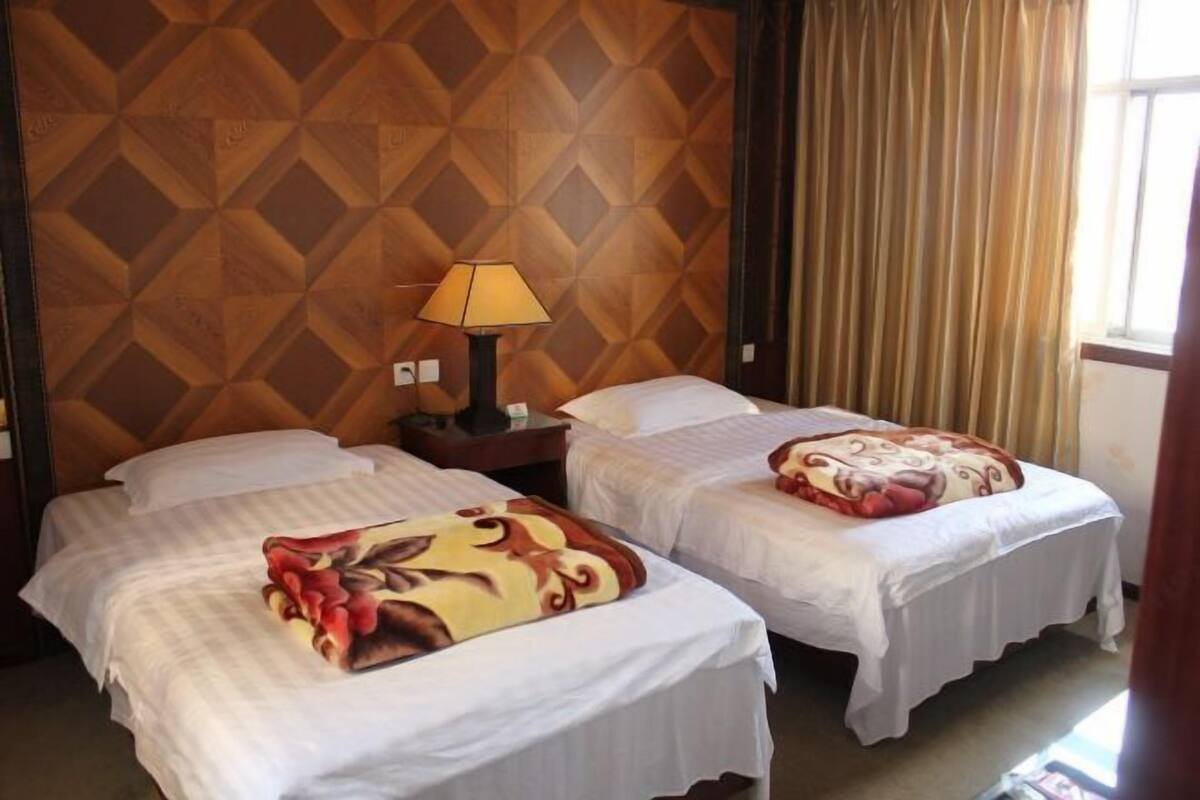 Ein Hotelzimmer mit zwei Betten, einem hölzernen Kopfteil, einem Nachttisch mit Lampe und einem Fenster mit Vorhängen.