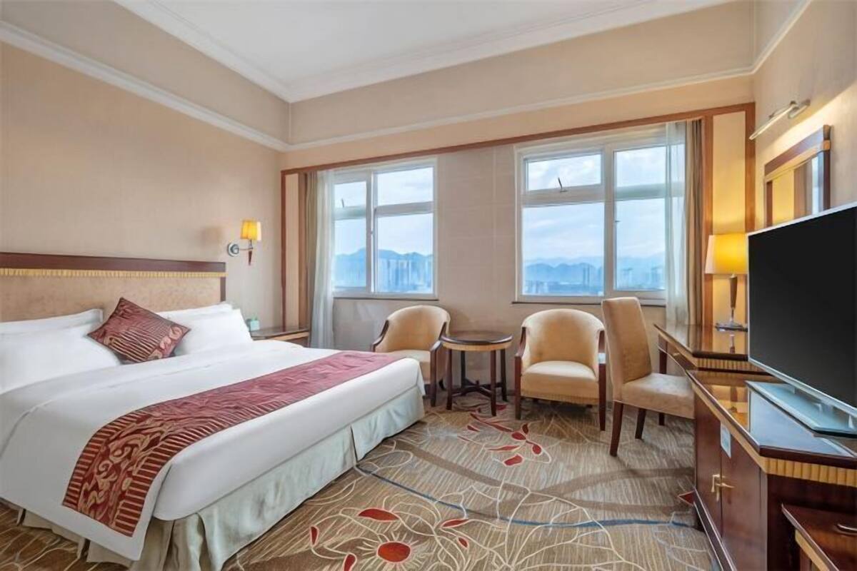 A hotel room with a large bed, two chairs, a TV, and a view of the city.