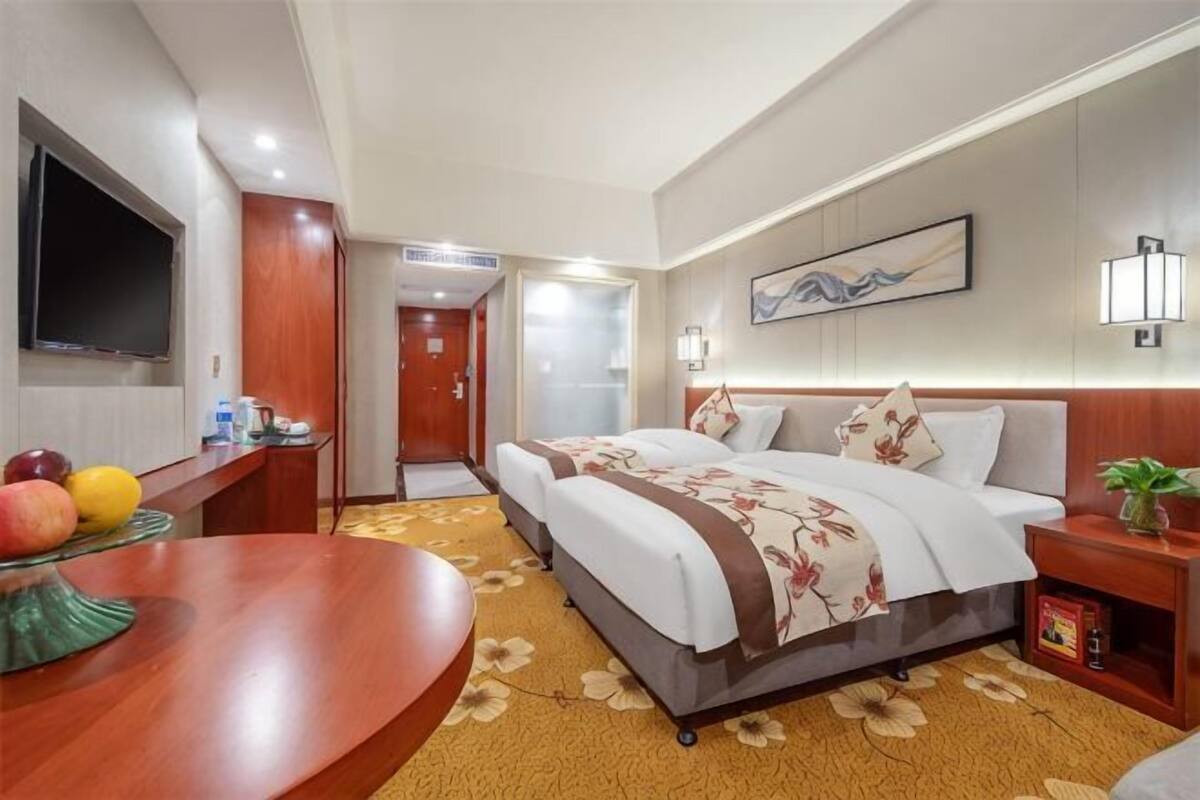 A hotel room with a bed, bedside table, TV, and a desk with fruits.