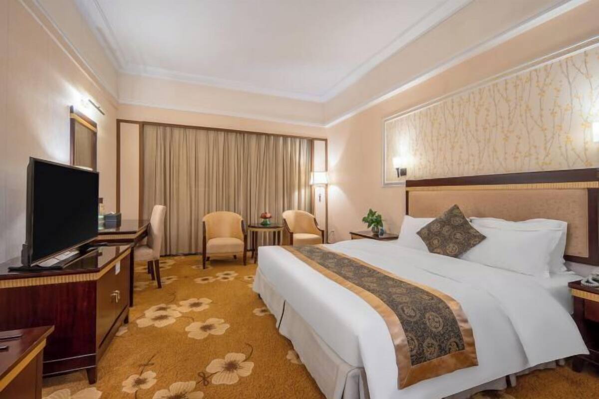 A hotel room with a bed, a desk with a TV, two chairs, a small table with a lamp, and a large window with curtains.