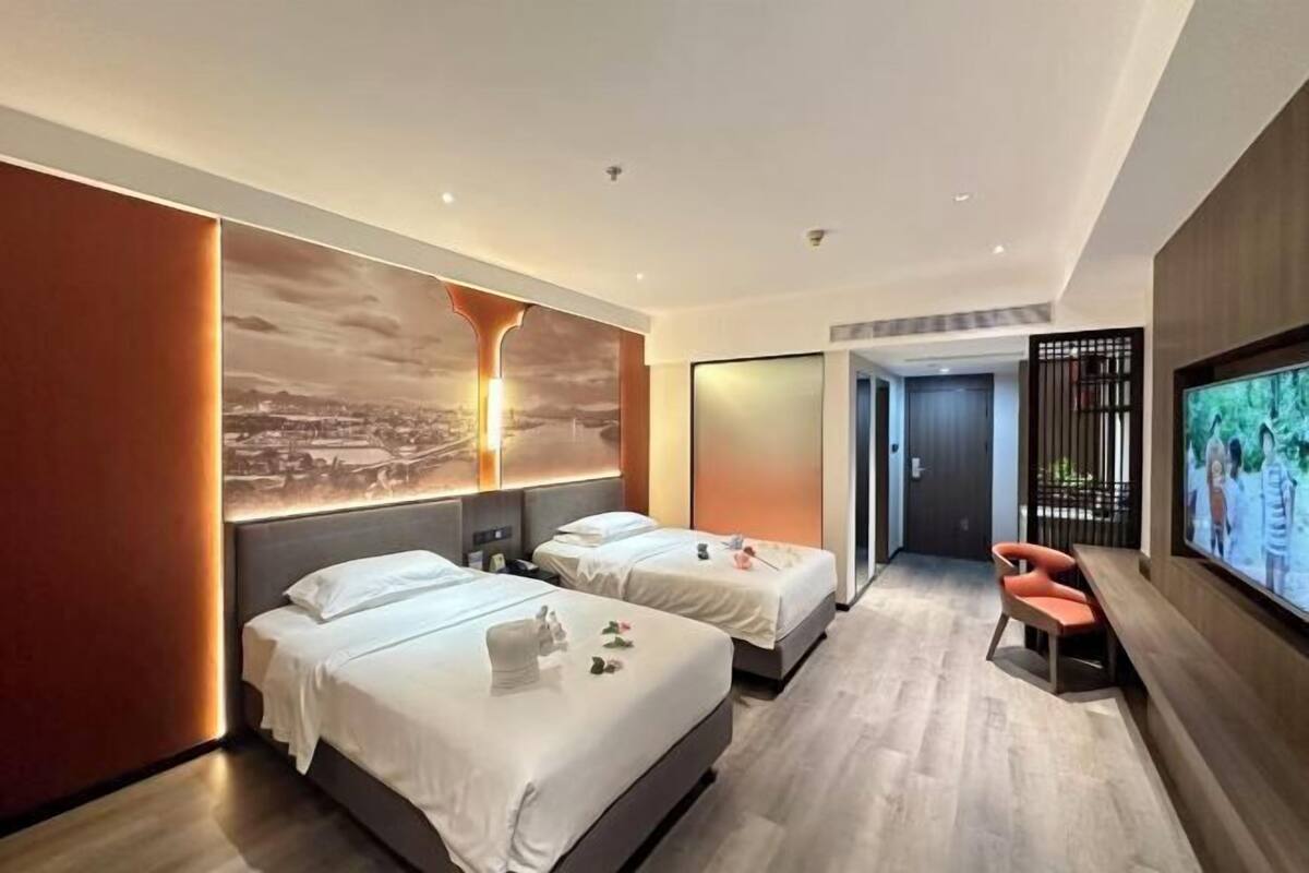 A modern hotel room with two beds, a large wall-mounted TV, and a wooden floor.