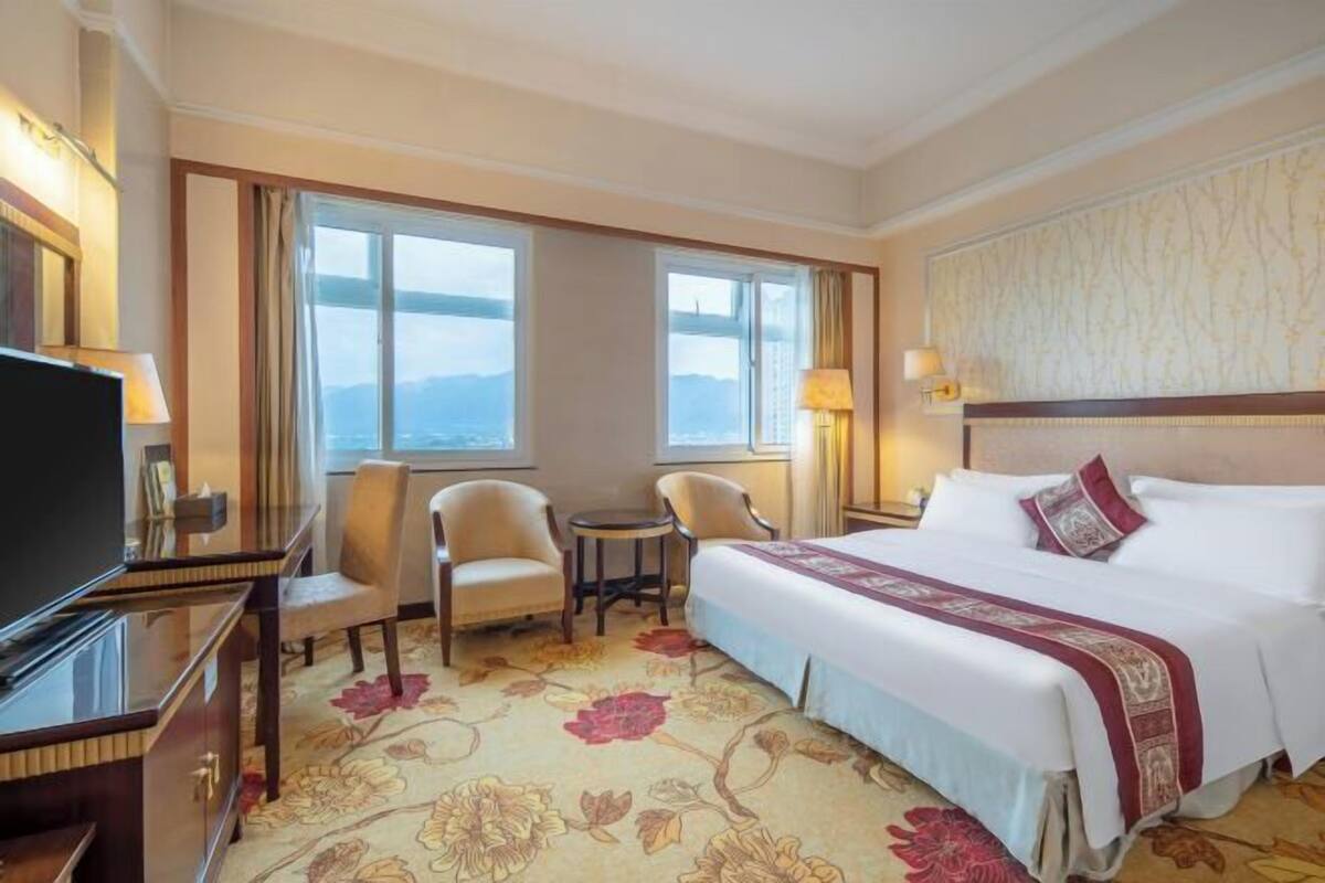 A hotel room with a large bed, two armchairs, a small table, a TV, and a view of the mountains through the windows.