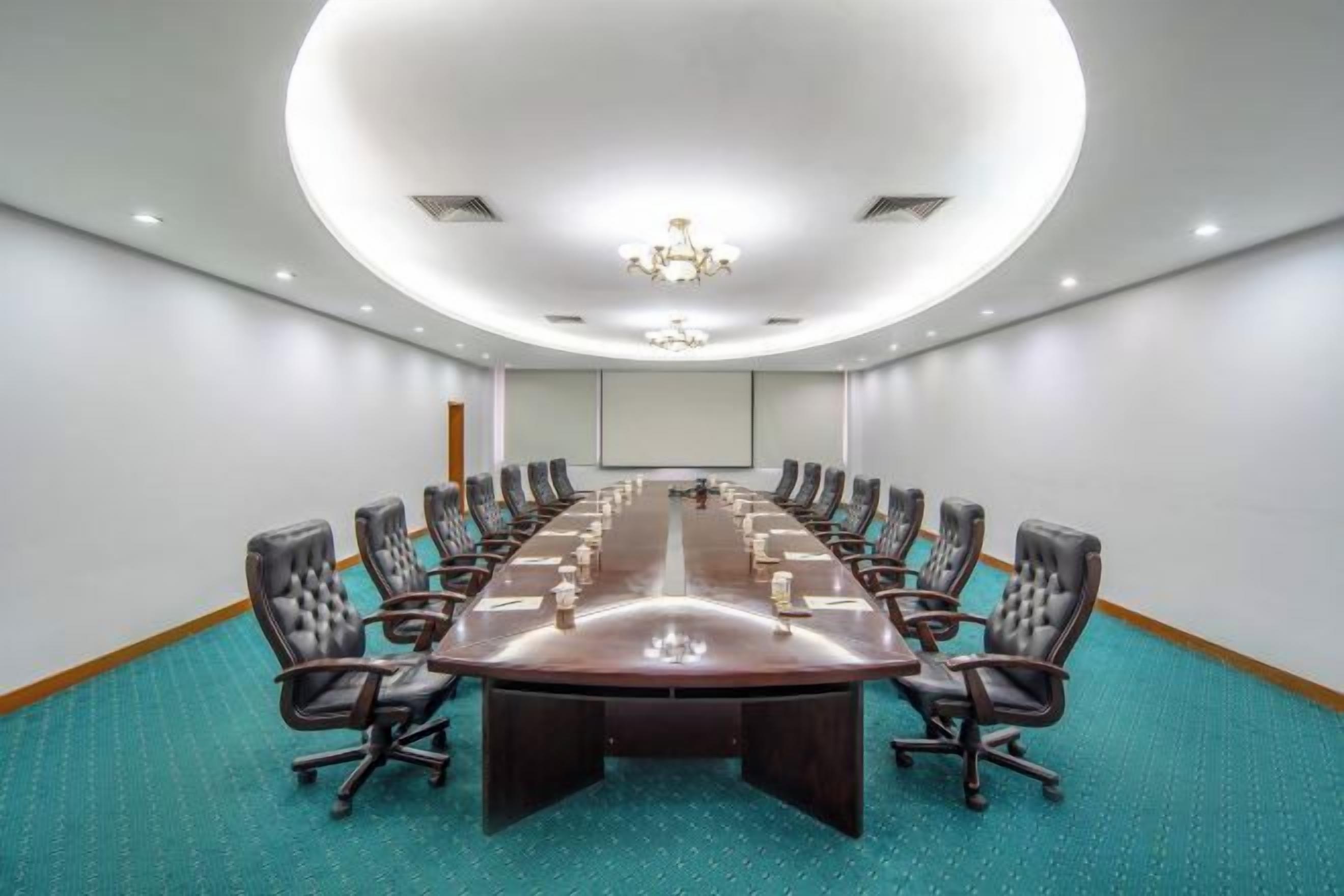 A conference room with a long table, multiple chairs, a projector screen, and a carpeted floor.