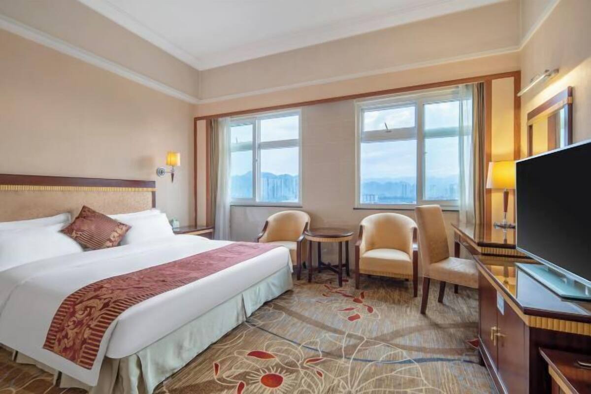 A hotel room with a large bed, a desk, two chairs, a television, and a view of the city through large windows.