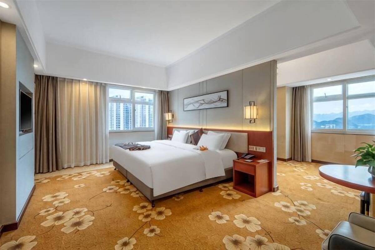 A hotel room with a large bed, a desk, a chair, a TV, and a view of the city.
