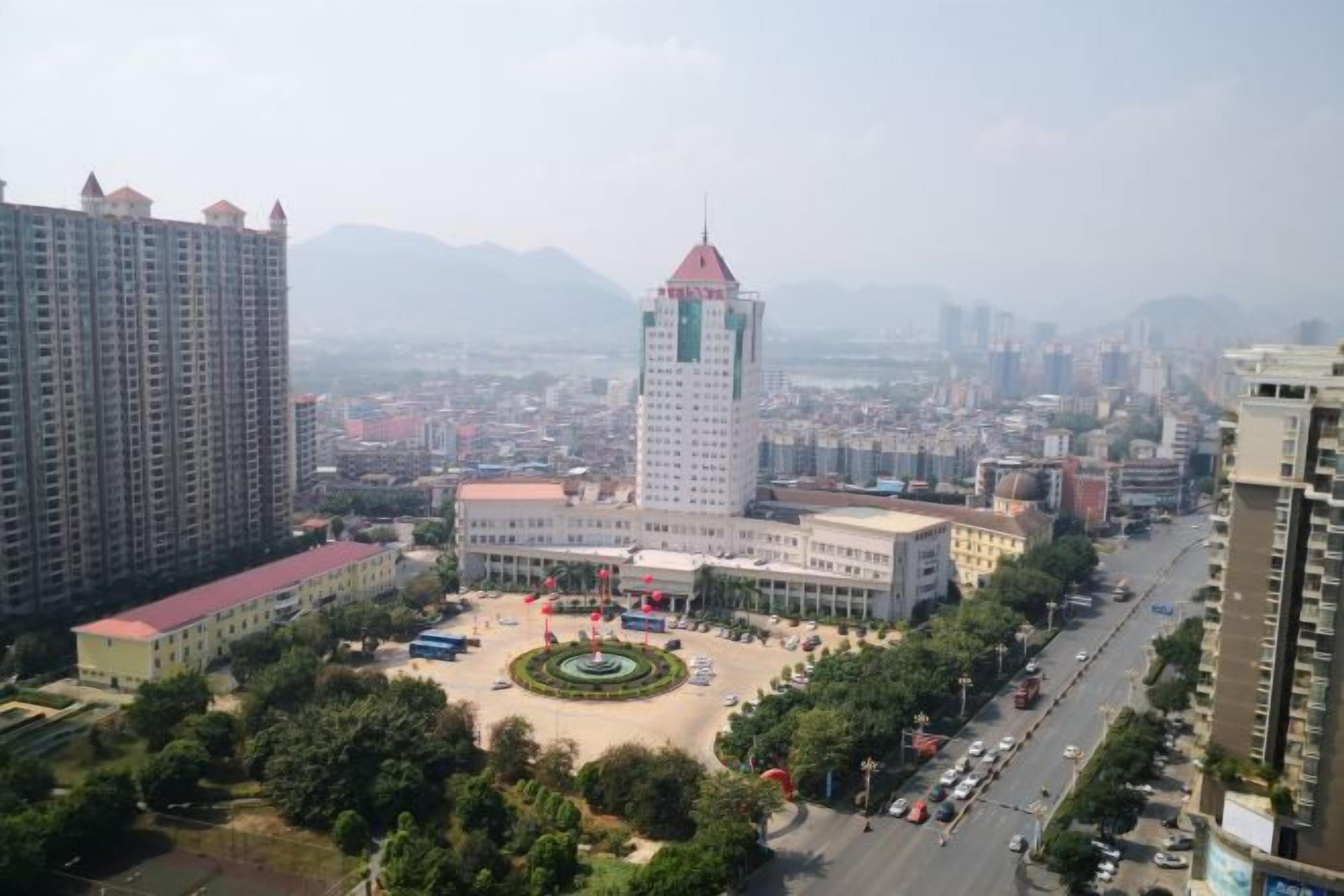 A cityscape with a prominent building with a red roof, surrounded by other high-rise buildings, a central plaza with a fountain, and a road with vehicles.