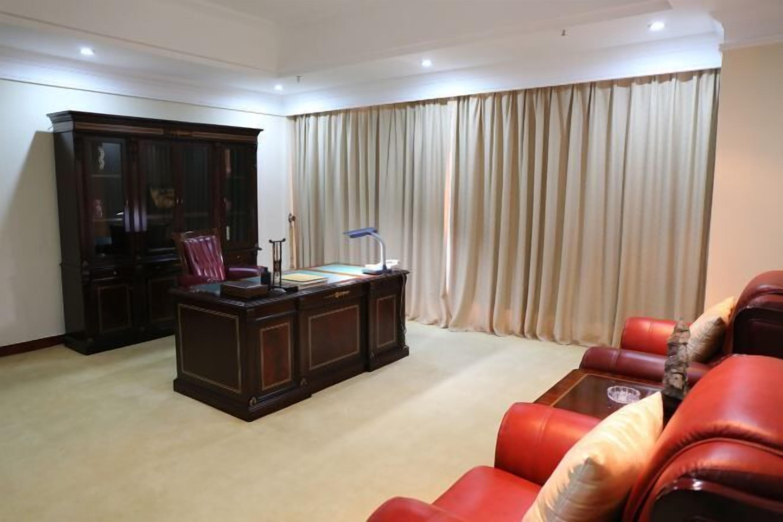 A room with a dark wooden desk, red leather sofa, and a large window with beige curtains.
