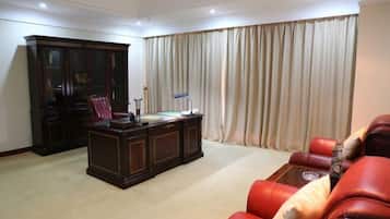 A room with a dark wooden desk, red leather sofa, and a large window with beige curtains.