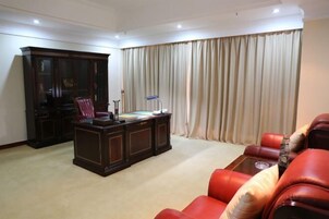 A room with a dark wooden desk, red leather sofa, and a large window with beige curtains.