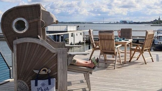 A deck with wicker chairs and a table, overlooking a body of water with a distant bridge. - Houseboat Liliput Comfortable Holiday Residence (Stralsund)