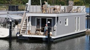 A floating houseboat docked at a marina with a covered outdoor seating area and a staircase leading to the upper deck. - Houseboat Liliput Comfortable Holiday Residence (Stralsund)