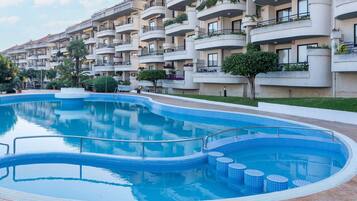 A modern apartment building with a central swimming pool, balconies with plants, and a well-maintained lawn.