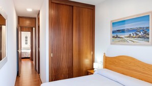 Une chambre d’hôtel avec un lit, une armoire en bois, une table de chevet avec une lampe et un tableau représentant un paysage côtier accroché au mur.